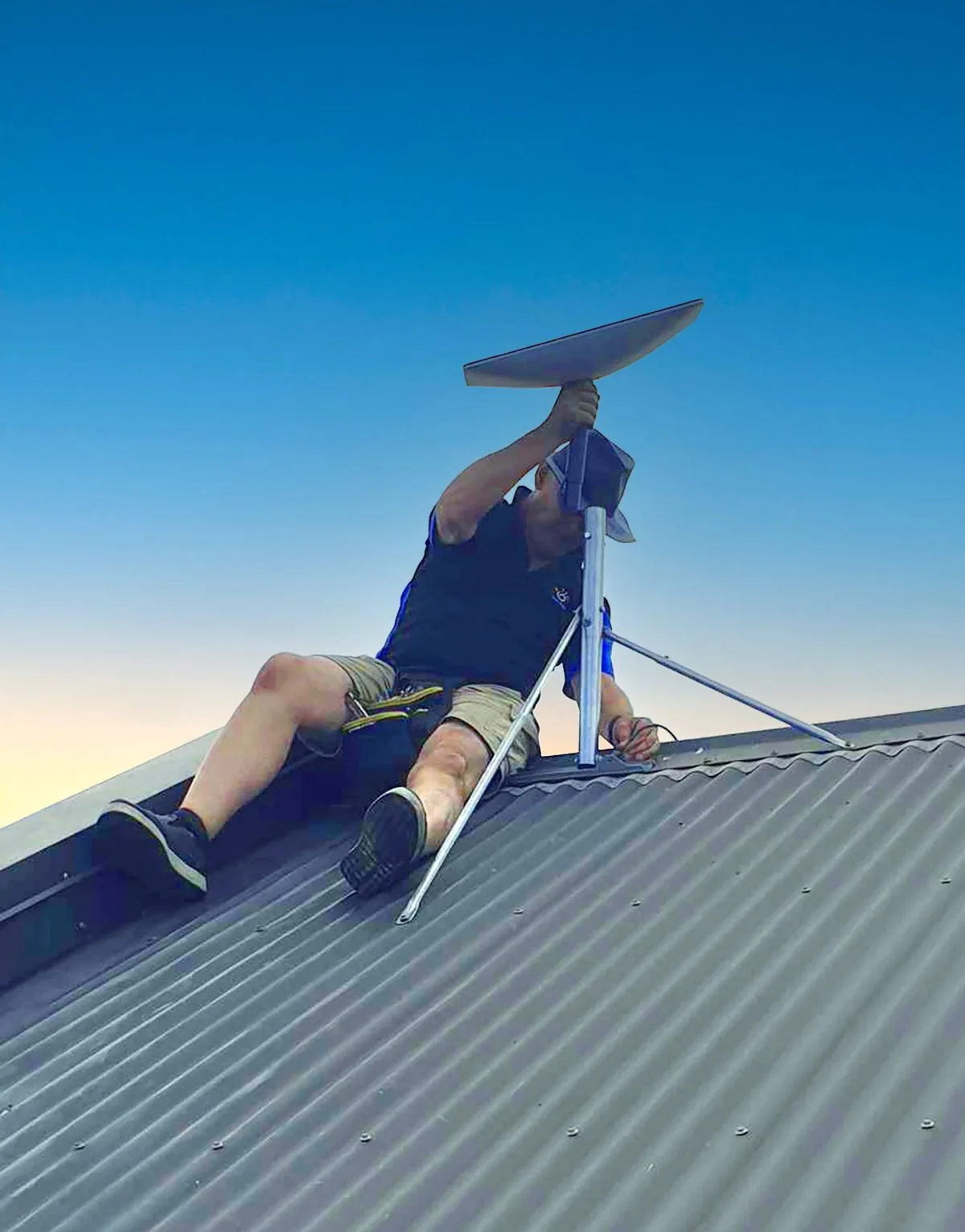 Glenn from Need Help NZ fitting a Starlink dish to a pole mount on a corrugated iron roof at dusk